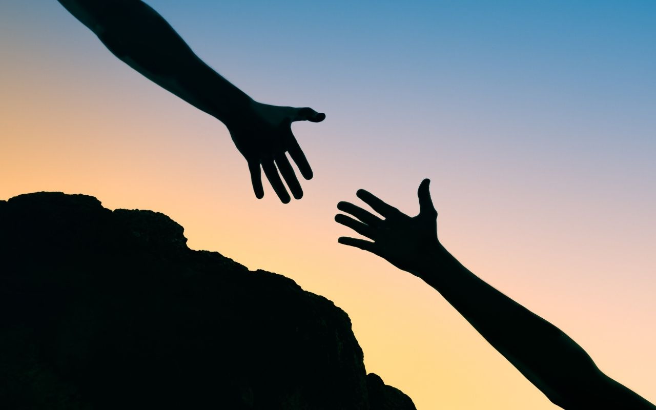 Two hands reaching for each other. One being lower and the other one reaching from above a mountain. On the back, there is a sunset.